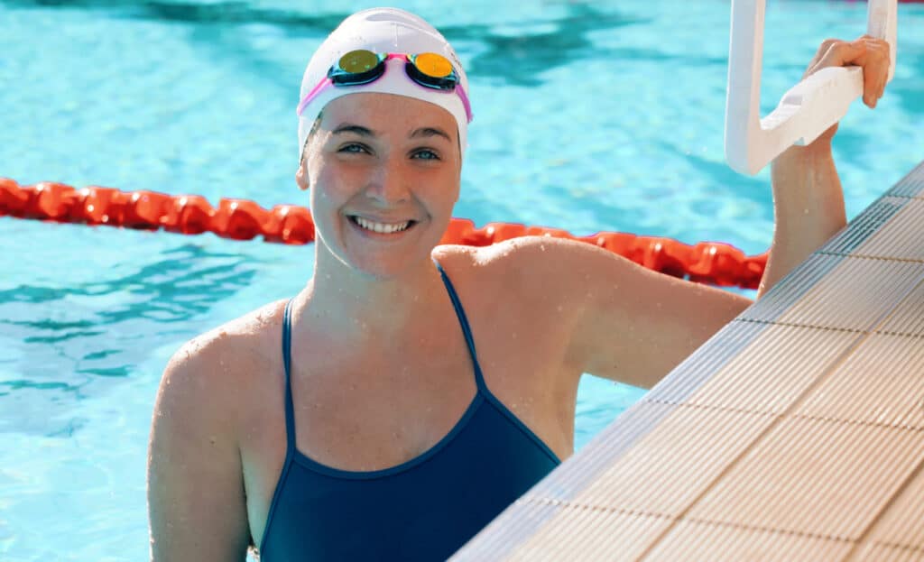 A young smiling woman in swimming costume, cap and goggles holds on to the edge of an Olympic swimming pool